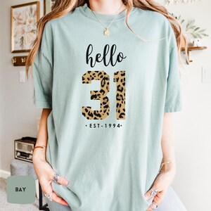 Puede incluir: Una camiseta azul claro con el texto "hello 31 - EST. 1994" impreso en ella. El número 31 está impreso en un patrón de estampado de leopardo.