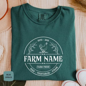 Puede incluir: Una camiseta verde azulada con un logotipo circular blanco que dice "ESTD - 2025" y "FARM NAME" con una ilustración de pollo dentro del círculo. El texto debajo del círculo dice "FARM FRESH" y "EGGS - VEGETABLES - STRAW".