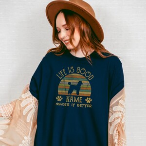 May include: A navy blue t-shirt with a vintage-style sunset graphic featuring a poodle silhouette. The text "Life is good [Name] makes it better" is printed on the shirt.