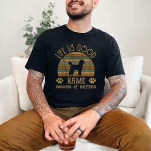 May include: A black t-shirt with a vintage-style sunset graphic and the text "Life is good [Name] makes it better". A silhouette of a poodle is in front of the sunset.