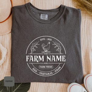 May include: A dark gray t-shirt with a white graphic design of a farm logo. The logo features a chicken, the text "FARM NAME", and the words "ESTD - 2025", "FARM FRESH", "EGGS", "VEGETABLES", and "STRAW".