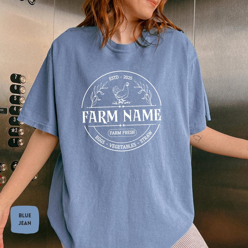 May include: A blue jean t-shirt with a white graphic of a farm logo. The logo includes a chicken, the text "ESTD - 2025", "FARM NAME", "FARM FRESH", and "EGGS - VEGETABLES - STRAW".