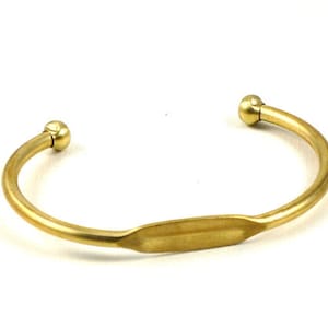 May include: A gold-colored cuff bracelet with a rounded, open design. The bracelet features a central, elongated oval shape and ball-shaped ends. The metal has a polished finish, reflecting light.
