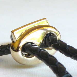 May include: A close-up of a gold-toned metal clasp with two black leather cords threaded through it.