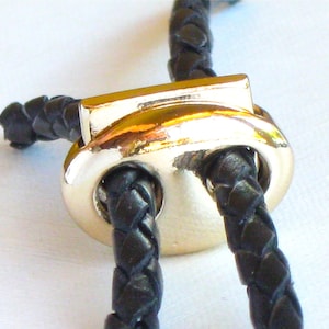 May include: Close-up of a gold-toned metal cord toggle with two holes for threading black braided leather cords.