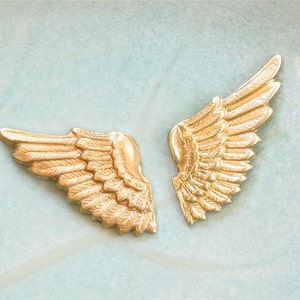 May include: Two gold-colored angel wings with detailed feather designs. The wings are separated and facing opposite directions.