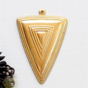 May include: Gold-colored triangular pendant with a textured surface and a small loop at the top. The pendant features a series of nested triangles, with a rope-like border. The pendant is likely for jewelry making.