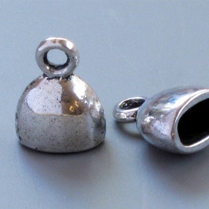 May include: Two silver-colored bell-shaped jewelry findings. Each finding has a loop at the top for attaching to a chain or other component. One finding is upright, and the other is on its side, showing the hollow interior.