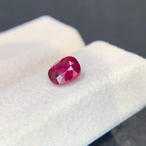 May include: A faceted, oval-shaped ruby gemstone. The gem is a deep, saturated red color, with visible internal facets that catch the light. It rests on a white, textured surface, with a dark background.