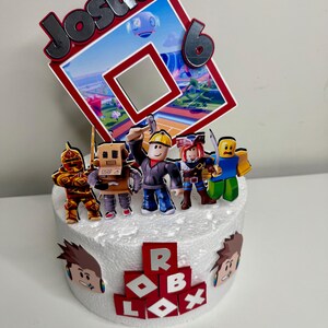 Puede incluir: Una tarta de cumplea&ntilde;os decorada con figuras de Roblox y un letrero que dice "Josua 6". La tarta presenta un logotipo de Roblox rojo y blanco, recortes de personajes y una base blanca.