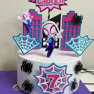 May include: A white cake decorated with a pink and blue spiderweb design, a pink number 7, a black spider, and a figure of Spider-Gwen. The cake is topped with a pink and blue sign that says "Lauren".