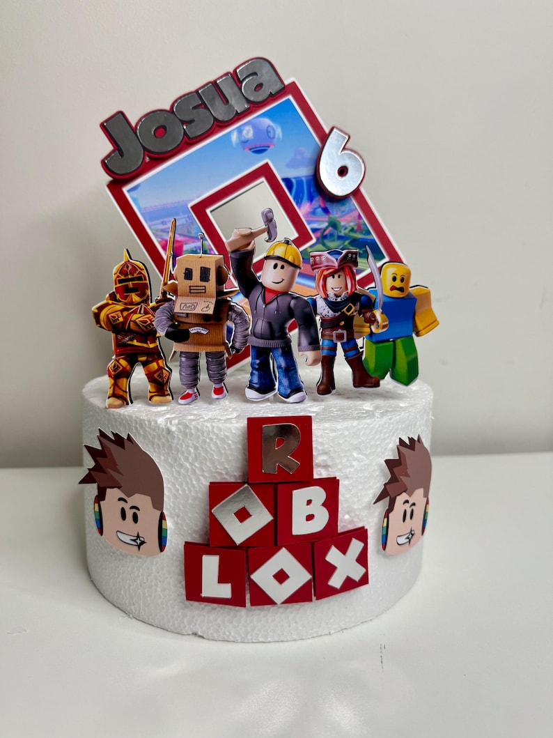 Puede incluir: Un adorno para pastel con tem&aacute;tica de Roblox con el nombre "Josua" y el n&uacute;mero "6". El pastel est&aacute; decorado con figuras de personajes de Roblox y el logotipo de Roblox. El pastel es blanco con detalles en rojo y plata.