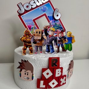 Puede incluir: Una tarta redonda blanca decorada con figuras de Roblox y un adorno que dice "Josua 6". La tarta presenta varios personajes de Roblox y el logotipo de Roblox. La tarta est&aacute; sobre una superficie blanca.