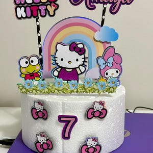 May include: A white cake decorated with Hello Kitty and My Melody characters, a rainbow, and a cloud. The cake is topped with a pink number 7 and the name "Amaya" in purple glitter letters.