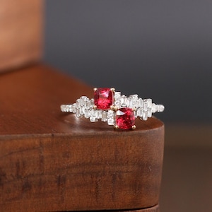 May include: A gold ring with two red gemstones and a band of small diamonds. The ring is sitting on a wooden box.