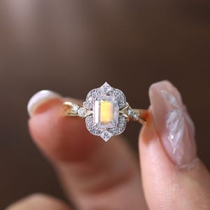 May include: A gold ring with a rectangular, light-colored gemstone at the center, surrounded by smaller, clear stones. The ring has a delicate, vintage design with intricate detailing on the band and around the main stone. The ring is held up by a person's fingers.