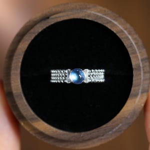 Puede incluir: Un anillo de plata con un cabujón central redondo de piedra lunar azul translúcida. La banda está compuesta por tres filas paralelas de pequeñas cuentas de plata texturizadas, creando un aspecto detallado de cuentas en toda la circunferencia.