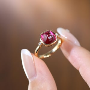 Könnte beinhalten: Ein Goldring mit einem zentralen, kissenförmigen, tiefrosa Edelstein in einer Zargenfassung, flankiert von zwei kleinen, runden, klaren Akzentsteinen auf beiden Seiten der Ringschiene. Das Metall hat ein poliertes Finish und einen warmen Goldton.