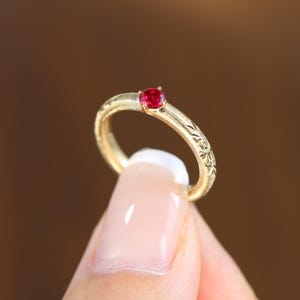 May include: A gold ring featuring a single round, vibrant red gemstone set in a prong setting. The band has a brushed, textured finish and is decorated with an engraved leaf or floral pattern along the sides of the metal shank.