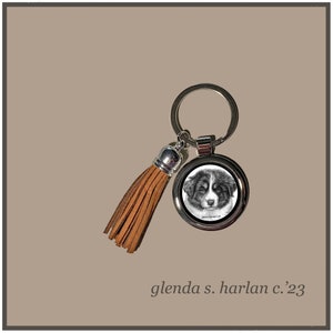 May include: A silver key chain with a brown leather tassel and a round charm featuring a black and white drawing of a Bernese Mountain Dog puppy. The charm has the text "Glenda S. Harlan '23" printed on it.