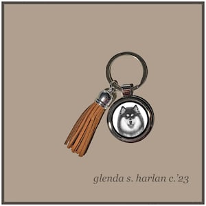 May include: A silver keychain with a brown leather tassel. The keychain features a round metal charm with a black and white image of a Finnish Lapphund dog.