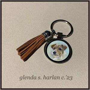Puede incluir: Un llavero plateado con un borla de gamuza marr&oacute;n y un dije redondo de metal con un retrato pintado a mano de un Jack Russell Terrier. El dije tiene un borde plateado y un fondo blanco. El texto "glenda s. harlan c.'23" est&aacute; impreso debajo de la imagen.
