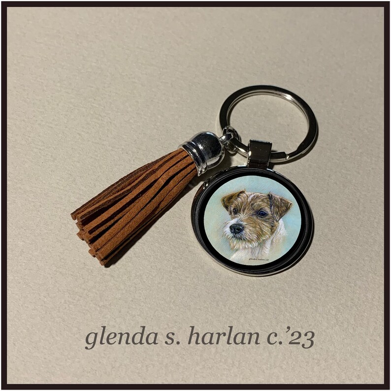Puede incluir: Un llavero plateado con un borla de gamuza marr&oacute;n y un dije redondo de metal con un retrato pintado a mano de un Jack Russell Terrier. El dije tiene un borde plateado y un fondo blanco. El texto "glenda s. harlan c.'23" est&aacute; impreso debajo de la imagen.