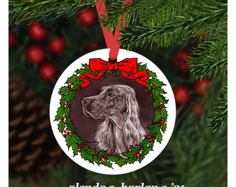 Könnte beinhalten: Ein runder weißer Weihnachtsbaumschmuck mit einem roten Band und einem grünen Stechpalmenkranz. Ein brauner und weißer Hundekopf befindet sich in der Mitte des Kranzes. Der Text "glenda s. harlan c. '21" befindet sich am unteren Rand des Schmucks.