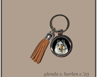 May include: A silver keychain with a brown leather tassel and a round metal charm featuring a portrait of a Collie dog. The charm has a black background and the dog's fur is white and black.