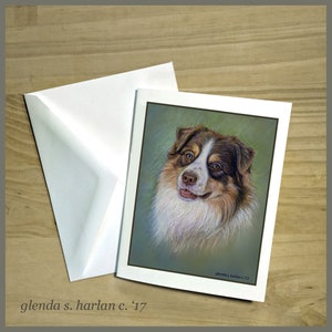 May include: A greeting card featuring a pastel drawing of an Australian Shepherd dog with brown, white, and black fur. The dog is looking directly at the viewer with a friendly expression. The background is a soft green color. The card is signed "glenda s. harlan c. '17".