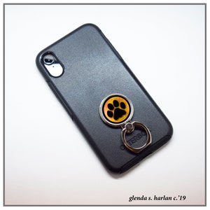 K&ouml;nnte beinhalten: Eine schwarze Handyh&uuml;lle mit einem silbernen Handyringhalter. Der Ringhalter hat ein gelbes und oranges Pfotenabdruck-Design.