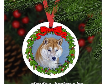 Könnte beinhalten: Ein runder weißer Weihnachtsbaumschmuck mit einem roten Band und einem grünen Stechpalmenkranz. Im Kranz befindet sich ein Gemälde eines Shiba Inu Hundes mit einem weißen und braunen Fell.
