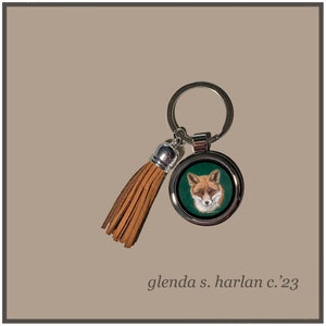 May include: A silver key chain with a brown leather tassel. The keychain features a round metal charm with a picture of a red fox on a green background. The text "glenda s. harlan c.'23" is printed below the image.