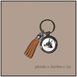 May include: A silver keychain with a brown leather tassel features a round, silver charm with a black and white illustration of a dog. The charm has the text "Glenda Harlan '23" printed on it.