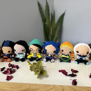 May include: A collection of eight crocheted dolls with different colored hair, clothing, and accessories. The dolls are arranged in a row on a white surface.