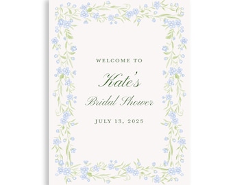 May include: A rectangular sign with a floral border of blue flowers and green leaves. The text reads "Welcome to Kate's Bridal Shower" and "July 13, 2025" in elegant script.