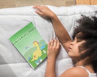 Puede incluir: Un cuaderno verde con la palabra alemana "HAUSAUFGABEN" y una ilustración de dinosaurio de dibujos animados. El cuaderno está sobre un edredón blanco junto a una persona.