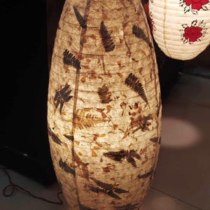 May include: A tall, oval-shaped paper lantern with a natural, earthy design. The lantern's surface is a light tan color, adorned with pressed leaves and fern patterns in shades of brown and black. A second lantern with red rose designs is visible in the background.
