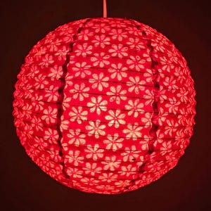 May include: A red paper lantern with a floral pattern. The round lantern is suspended from a white cord and emits a warm glow. The white floral pattern contrasts with the red background, creating a decorative lighting element.