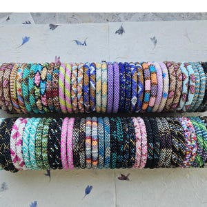 Puede incluir: Dos expositores cilíndricos negros que sostienen una colección de numerosas pulseras de cuentas. Las pulseras presentan una gran variedad de colores, incluyendo tonos de azul, rosa, verde, amarillo y negro, dispuestos en diversos patrones geométricos y florales.