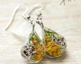 May include: Teardrop earrings with silver hooks, filled with yellow and green dried flowers set in clear resin. The earrings are displayed on a light wood surface, with a sprig of greenery in the background.