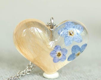 May include: A heart-shaped pendant necklace with a silver chain. The pendant is clear with a section of light brown material and blue forget-me-not flowers. The necklace is on a gray surface with white flowers in the background.