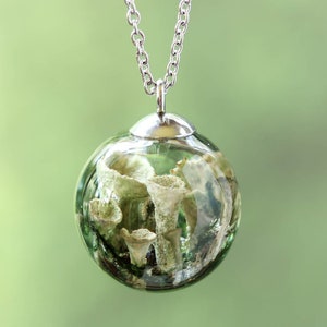 May include: A clear glass orb pendant with a silver chain. The orb contains a miniature landscape of green and white organic shapes, possibly resembling plants or fungi. The background is a soft, blurred green.