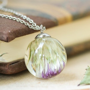 May include: A silver chain necklace with a clear glass pendant containing a preserved flower with green and purple hues.