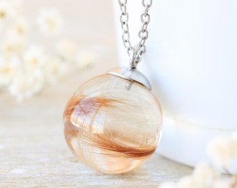 May include: A clear, spherical pendant necklace with a silver chain. The pendant features strands of brown hair encased in clear resin. The necklace is set against a white backdrop with small white flowers.