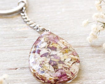 May include: A teardrop-shaped keyring featuring a clear resin pendant filled with small, dried pink and white flower petals. The pendant is attached to a silver-toned metal split ring by a short, delicate silver-toned metal chain link.