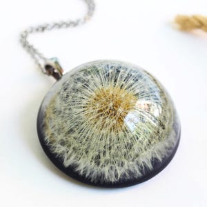 May include: A dandelion seed head pendant necklace. The round pendant is encased in clear resin with a dark blue border, suspended from a silver chain. Dried wheat stalks are arranged around the necklace on a white surface.