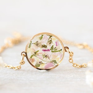 Op de afbeelding: Een goudkleurige ketting met een ronde hanger gevuld met gedroogde bloemen in roze, wit en groen. De fijne ketting ondersteunt de hanger, die een natuurlijk bloemenarrangement toont. De achtergrond is een zachte, neutrale kleur.