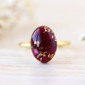 Op de afbeelding: Een gouden ring met een ovale, dieprode edelsteen met gouden vlokken. De ring staat op een licht houten oppervlak, met zachte, vage witte bloemen op de achtergrond.