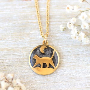 Pode incluir: Colar com pingente dourado apresentando a silhueta de um gato dentro de uma moldura circular. O gato é retratado em uma pose de caminhada contra um fundo escuro, com um detalhe de lua crescente. A corrente do colar também é dourada.